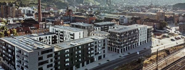 Luftbild einer Stadtansicht mit modernen Wohngebäuden im Vordergrund, einem alten Fabrikschornstein und Weinbergen im Hintergrund.