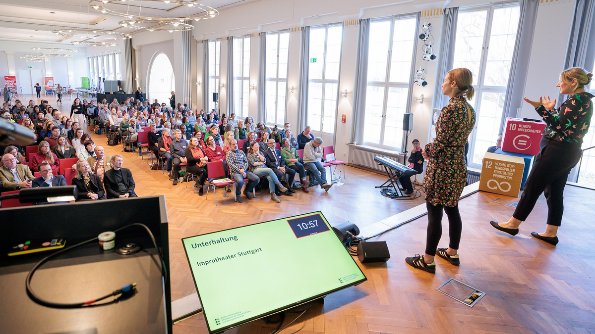 Auf dem Farbfoto stehen zwei Frauen auf einer Bühne und tragen etwas vor. Im Publikum sitzen Menschen auf roten Stühlen. Auf einem Bildschirm auf der Bühne steht „Unterhaltung, Improtheater Stuttgart“. Vorne rechts sitzt ein Pianist.