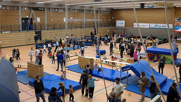 Turnhalle in der viele Menschen sind und verschiedene Stationen eines Parkours mit blauen Matten und Sprungelementen machen