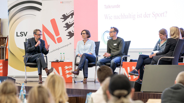 Farbfoto einer Podiumsdiskussion. Fünf Personen sitzen auf einer Bühne, eine Person spricht ins Mikrofon. Im Hintergrund eine Leinwand mit der Frage: „Wie nachhaltig ist der Sport?“