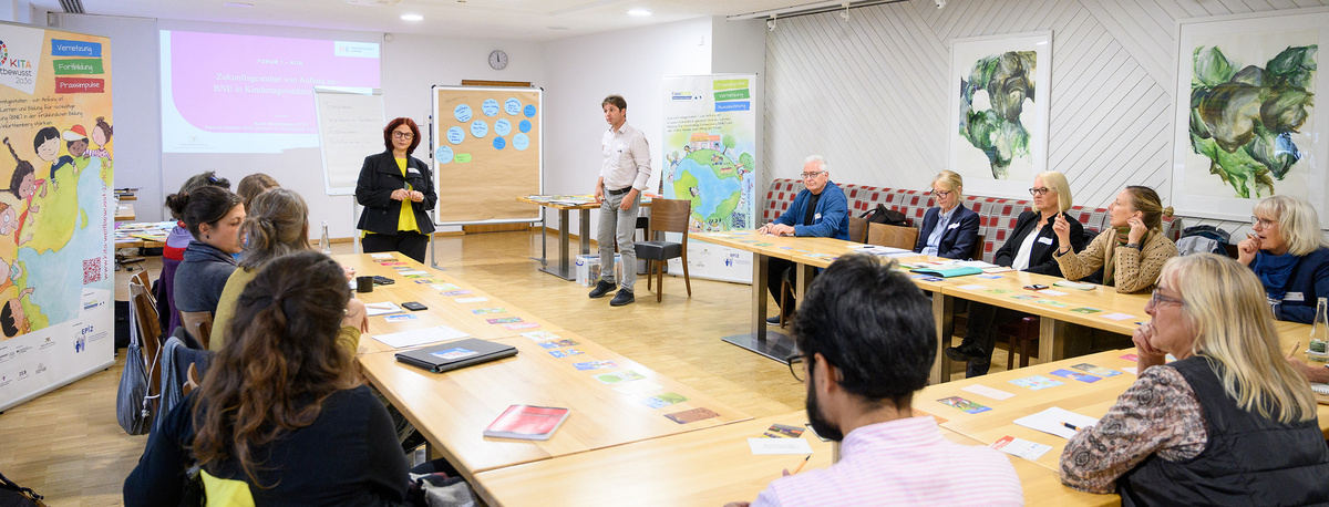 Mehrere Personen sitzen an U-förmig aufgestellten Tischen in einem Workshop, zwei Personen stehen vorne und richten den Blick auf die Menge.