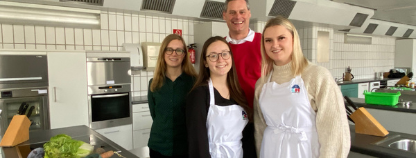 Gruppe von vier Personen in einer modernen Küche, zwei mit weißen Schürzen, die lächelnd vor einer Arbeitsfläche mit frischem Gemüse stehen.