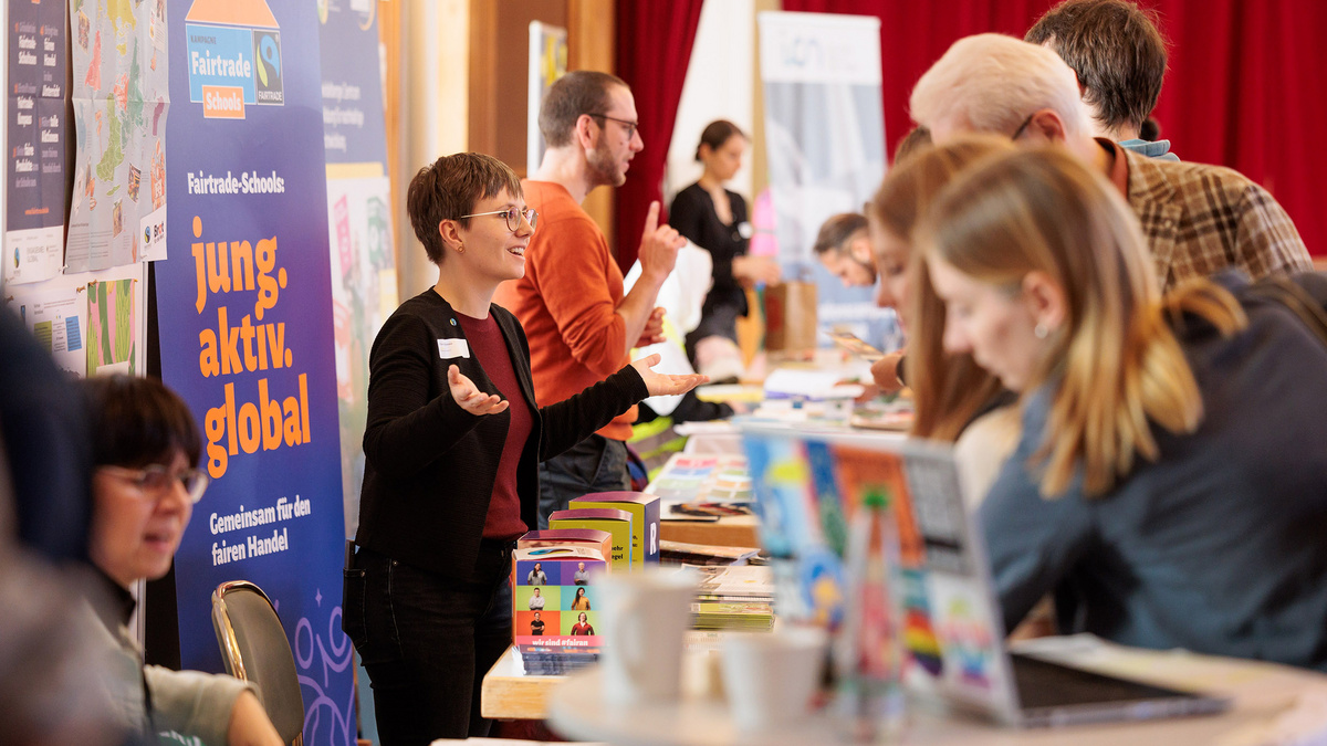 Das Farbfoto zeigt Menschen, die an Infoständen in einem Saal stehen. Eine Frau gestikuliert im Gespräch. Ein Plakat hinter ihr trägt den Text: „Fairtrade-Schools: jung. aktiv. global.“