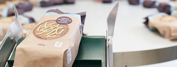 Ein Nudelpaket in Papierverpackung läuft von einem Förderband auf einen Tisch zur Weiterverarbeitung.