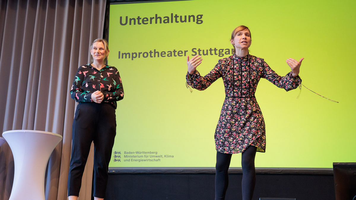 Auf dem Farbfoto stehen zwei Frauen mit Ansteckmikrofonen auf einer Bühne und tragen etwas mit Handgestiken vor. Auf einem Bildschirm auf der Bühne steht „Unterhaltung, Improtheater Stuttgart“. 