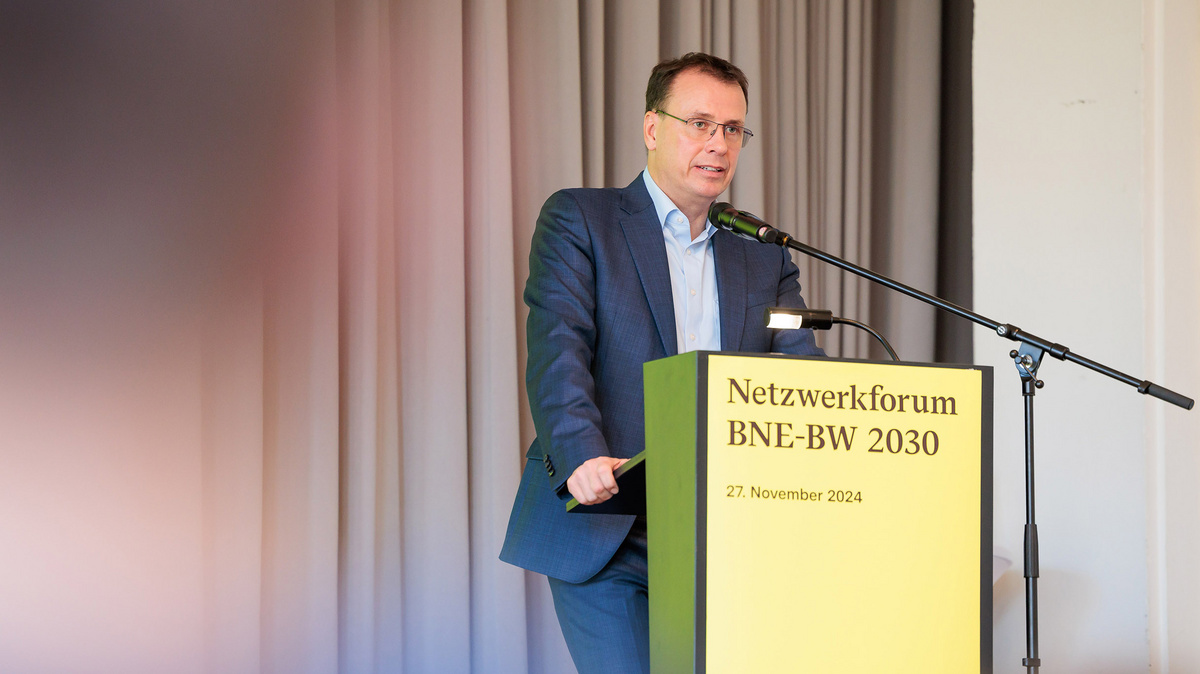 Der Kultusstaatssekretär Volker Schebesta MdL ist auf diesem Farbfoto auf einer Bühne am Rednerpult zu sehen. Darauf steht: „Netzwerkforum BNE-BW 2030, 27. November 2024“. Ein Mikrofon ist angebracht.