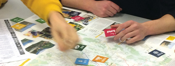 Auf dem Farbfoto sind die Hände zweier Personen über einer Landkarte, die auf einem Tisch liegt, zu sehen. Sie ordnen bunte, aus Papier ausgeschnittene Symbole auf der Karte an.