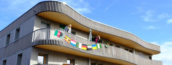 Holzverkleidetes, geschwungenes Mehrfamilienhaus mit Balkonen, geschmückt mit internationalen Flaggen, strahlender Himmel im Hintergrund.