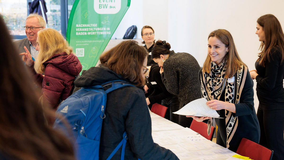 Menschen stehen an einem Tisch zur Akkreditierung. Im Hintergrund ein grünes Banner mit der Aufschrift „Green Event BW“. Eine Frau wendet sich an Teilnehmende.