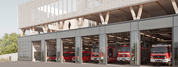 Moderne Feuerwehrstation mit großen Glasgaragen, durch die rote Einsatzfahrzeuge sichtbar sind, darüber eine Holzfassade.