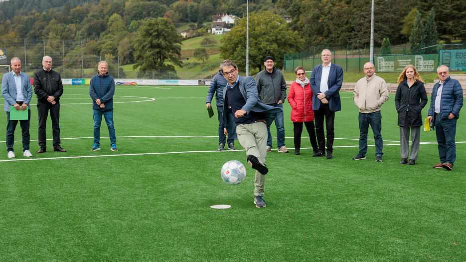 Staatssekretär Dr. Andre Baumann kickt einen Fußball über den Sportplatz in Heiligkreuzsteinach. 