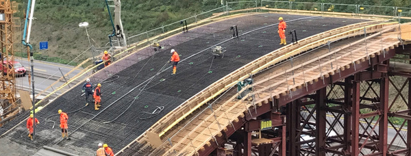 Baustelle einer Brücke mit mehreren Arbeitern in orangefarbener Schutzkleidung, Stahlgerüste und ein Betonpumpenarm sichtbar.